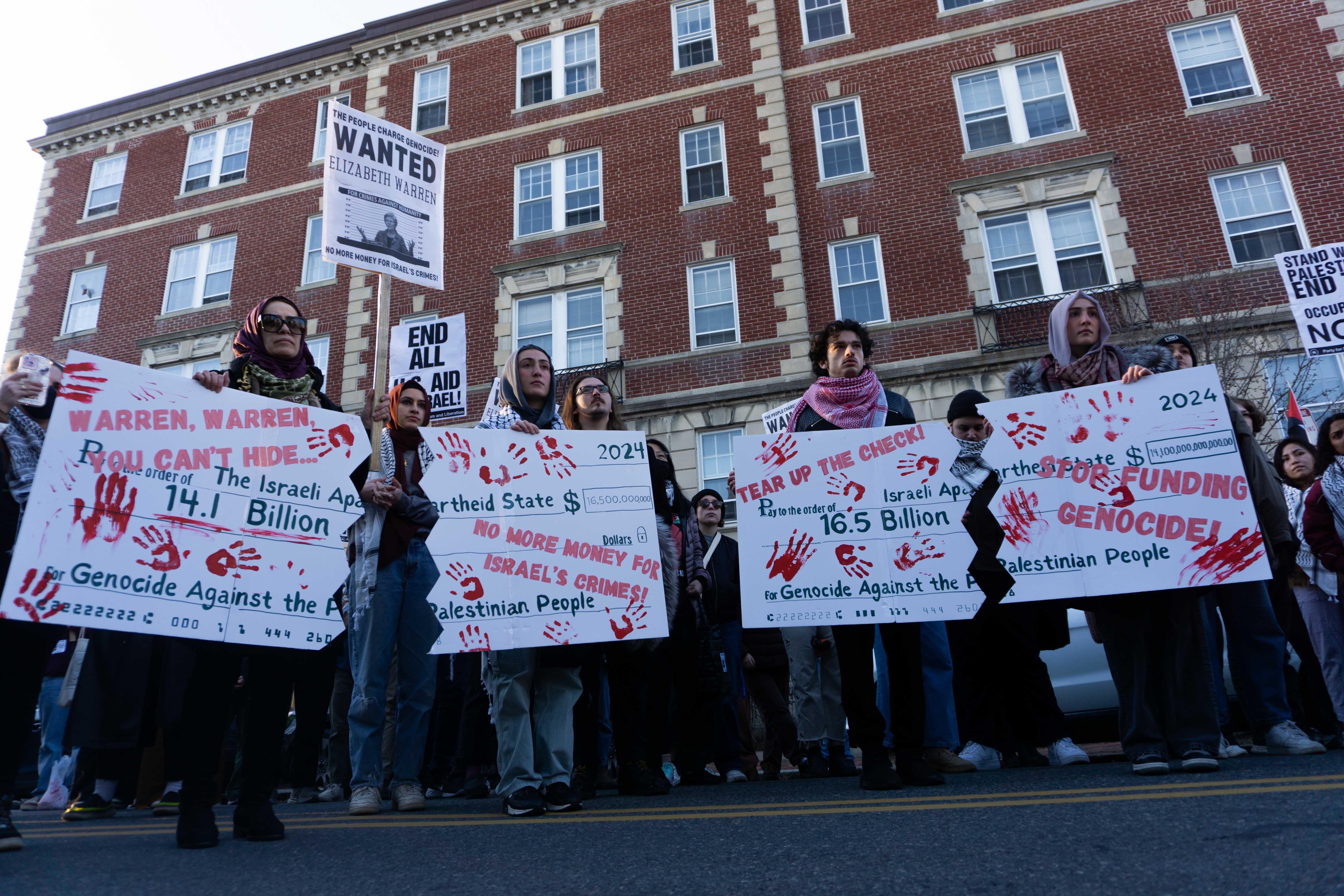 Protesters hold posters drawn as checks made out to “The Israeli Apartheid State” for “Genocide Against the Palestinian People” during a pro-Palestine rally. More than 200 people marched from Cambridge Common to the home of U.S. Sen. Elizabeth Warren (D-Mass.), demanding for her to vote against U.S. military aid to Israel.