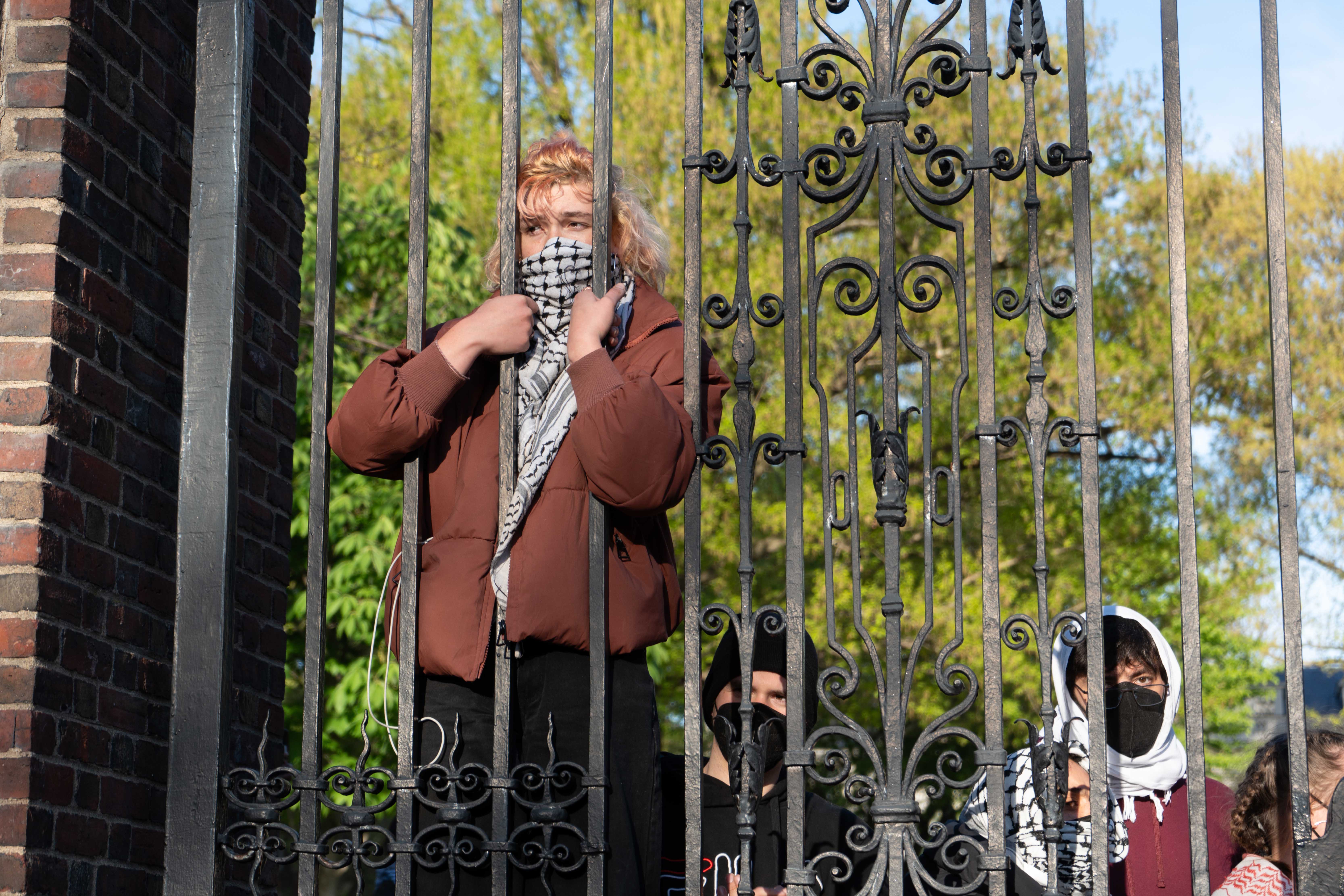 A pro-Palestine protester partially climbs Johnston Gate from within Harvard Yard to watch more than 200 people rally outside the Yard. The rally came one day after Harvard administrators placed 20 students on involuntary leave for participating in the encampment.