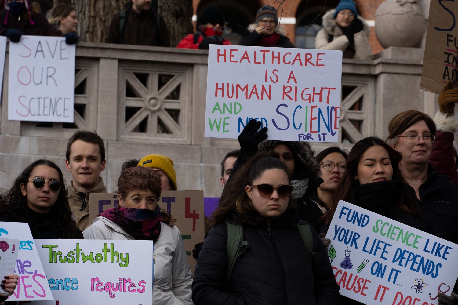Demonstrators hold signs on the Robert Gould Shaw Memorial in front of the Massachusetts State House during the Stand Up for Science rally protesting President Donald Trump’s proposed cuts to federal research funding. More than 500 researchers, physicians, and students gathered at Boston Common to oppose the administration’s policies.
