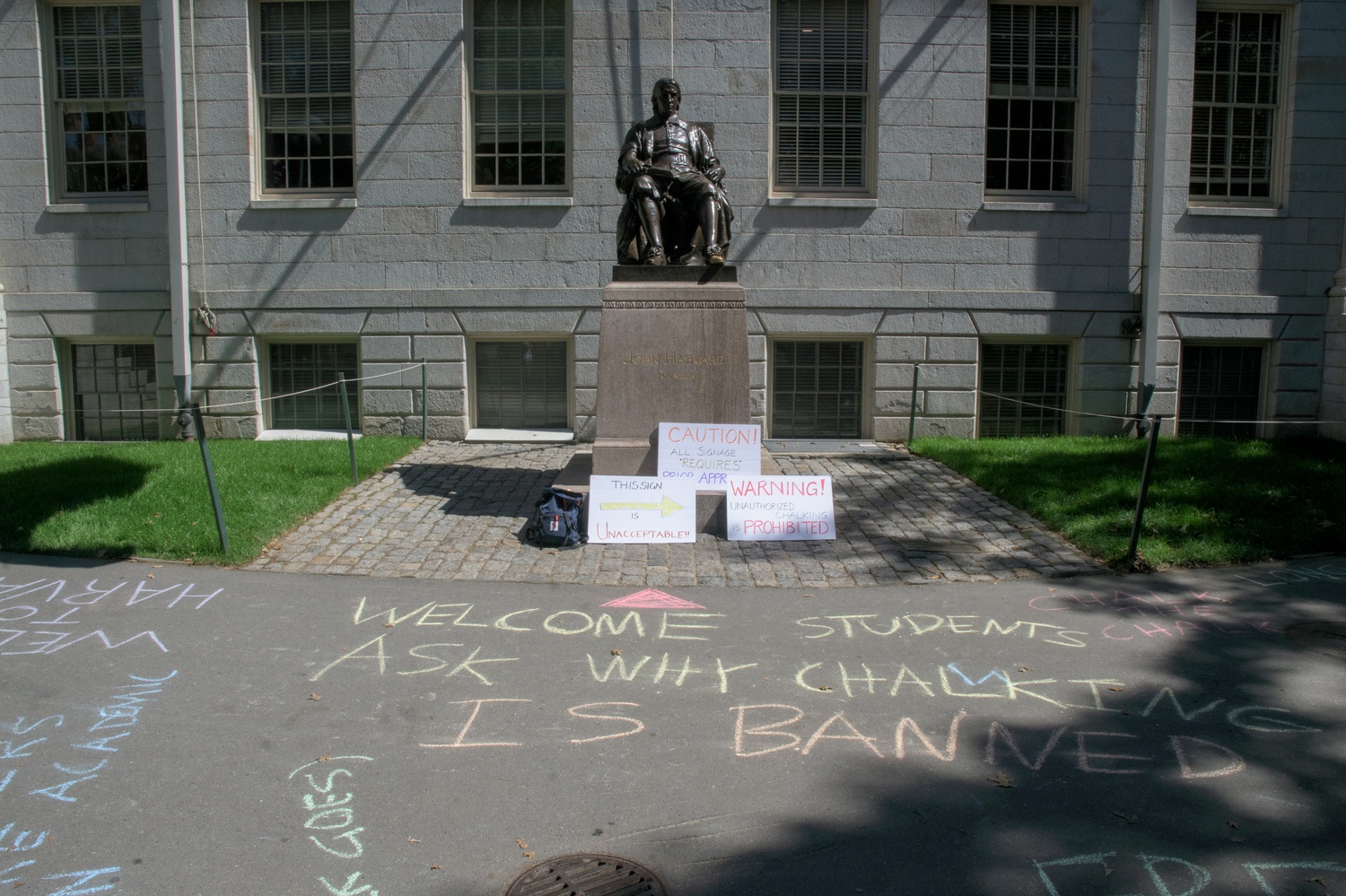 Messages in colorful chalk cover the sidewalk in front of the John Harvard statue during a faculty protest against new campus rules banning chalking and unsanctioned signage. One message reads, “Welcome students, ask why chalking is banned.” The chalk was washed away by Harvard staff less than an hour later.